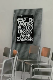 Black poster for Zagreb Design Week on a metallic grid wall behind empty silver metal chairs in a bright interior space.