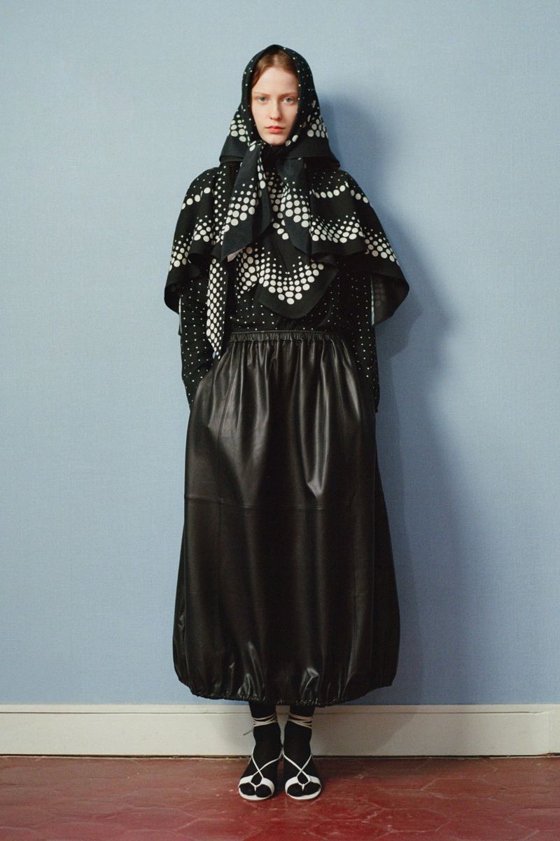Full-length fashion portrait of a woman with a headscarf, patterned top, leather skirt, socks, and shoes against a light blue wall.