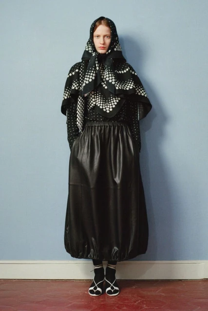 Full-length fashion portrait of a woman with a headscarf, patterned top, leather skirt, socks, and shoes against a light blue wall.