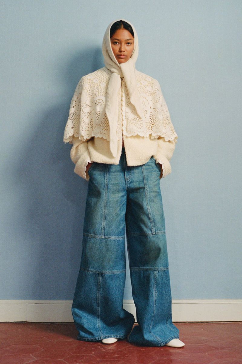 Fashion shot of a woman wearing a cream-colored lace cape with a hood, cream sweater and very wide-leg blue jeans standing indoors.