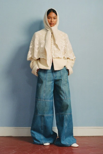 Fashion shot of a woman wearing a cream-colored lace cape with a hood, cream sweater and very wide-leg blue jeans standing indoors.