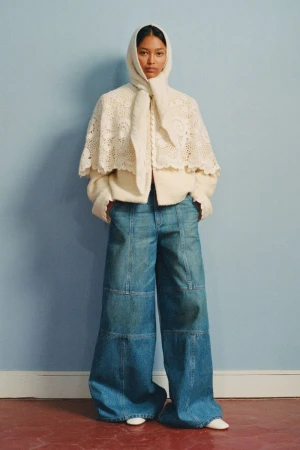 Fashion shot of a woman wearing a cream-colored lace cape with a hood, cream sweater and very wide-leg blue jeans standing indoors.