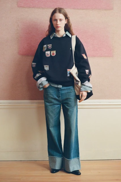 A long-haired model poses indoors wearing a dark crewneck sweatshirt with patches, blue jeans, and black shoes against a neutral backdrop.