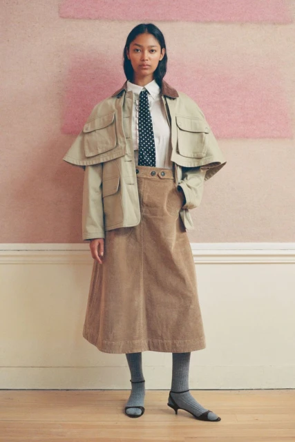 Fashionable woman in tan jacket, white shirt, and corduroy skirt poses in an indoor setting with hardwood floors and neutral-toned wall.