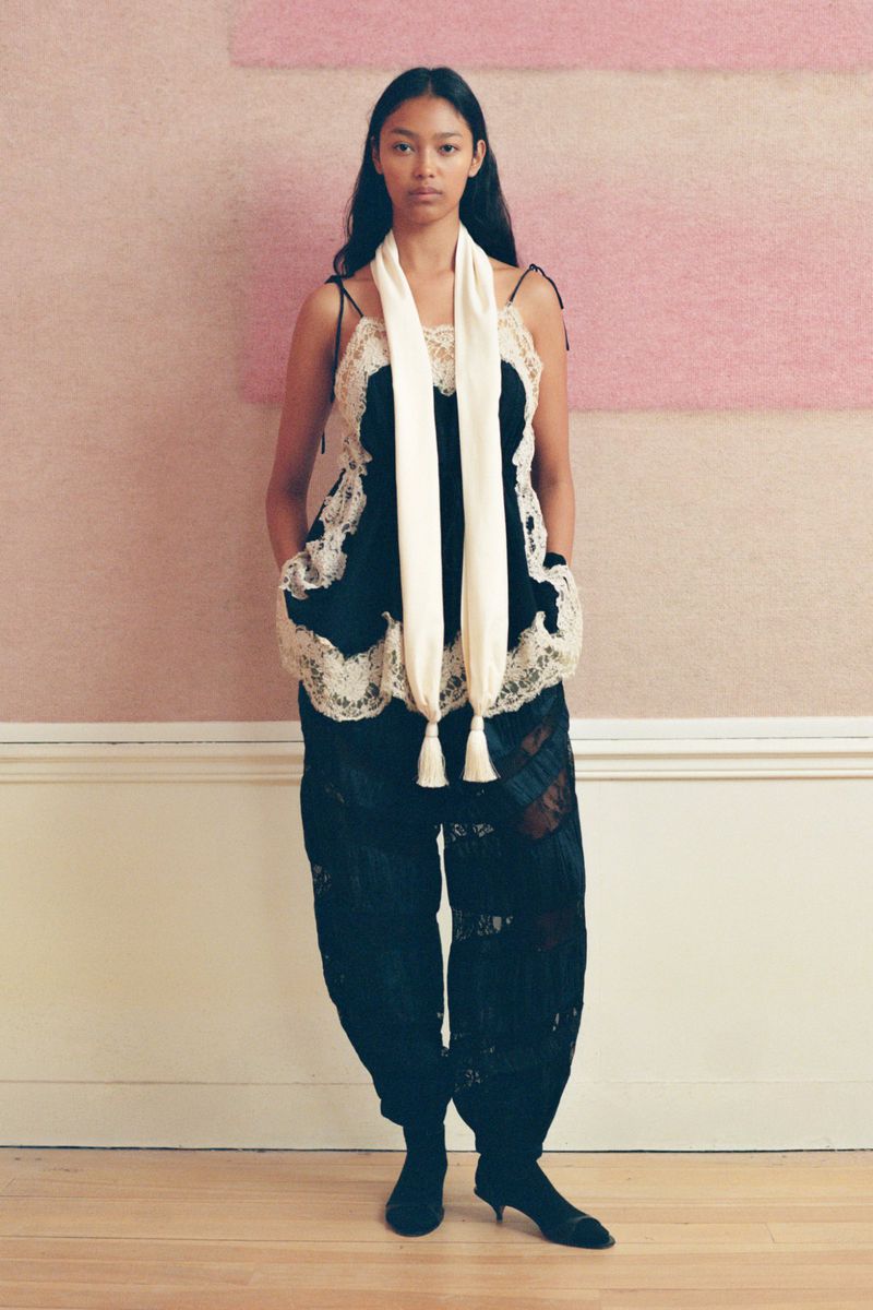 Adult woman posing indoors wearing a black lacy camisole top, black lacy pants, a white scarf, and black heeled shoes.
