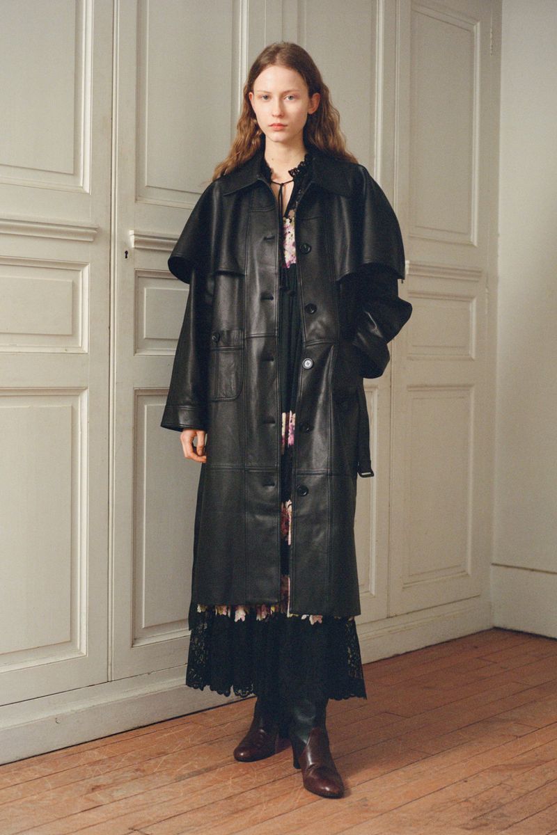 A fair-skinned young woman poses indoors, wearing a full-length black leather coat over a floral dress and brown leather boots.