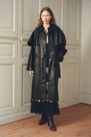 A fair-skinned young woman poses indoors, wearing a full-length black leather coat over a floral dress and brown leather boots.