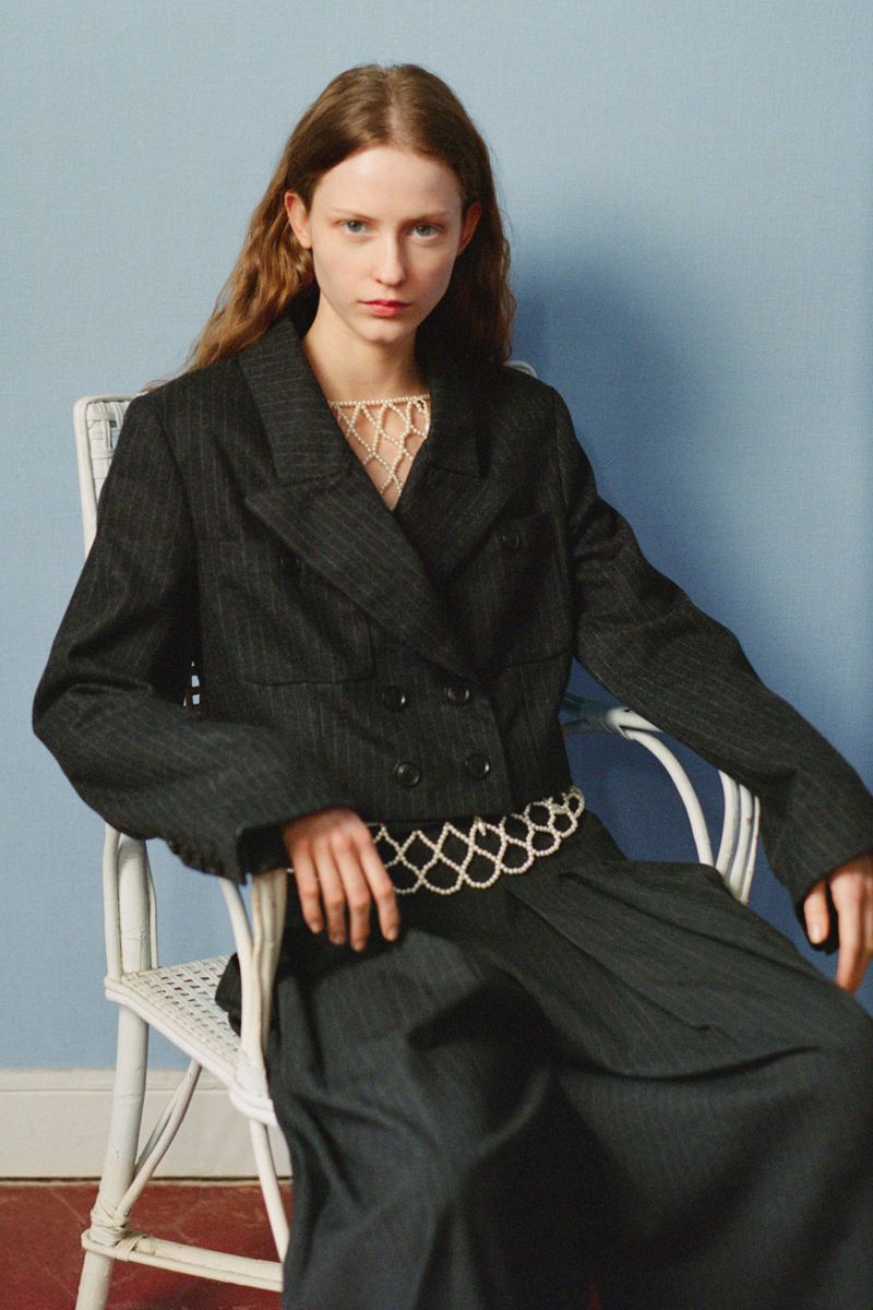 Portrait of a young adult woman with long hair wearing a dark jacket and pants, seated on a white chair against a blue wall.
