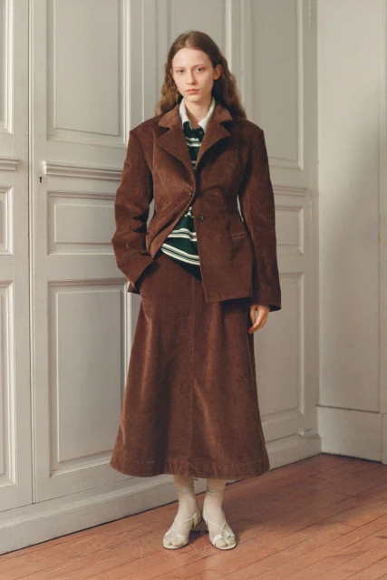 Adult female fashion model poses in brown corduroy blazer and matching skirt in an indoor setting with a neutral background.