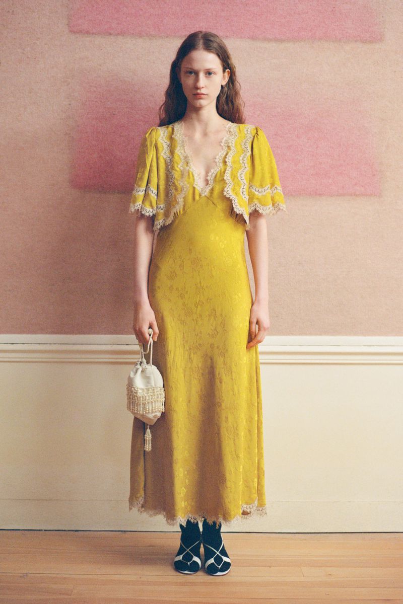 Elegant woman in mustard yellow floral dress with lace trim holding a beaded purse. Indoor setting with soft pink wall.