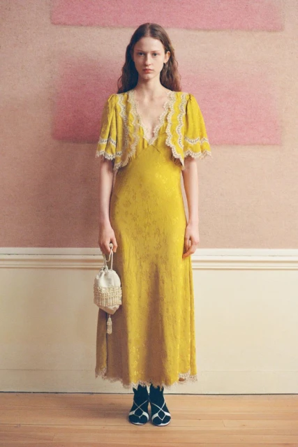 Elegant woman in mustard yellow floral dress with lace trim holding a beaded purse. Indoor setting with soft pink wall.