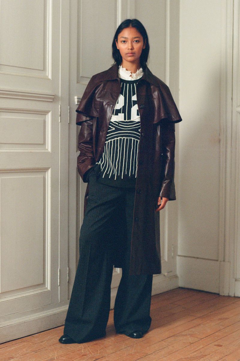 A stylish woman poses in a long brown coat, patterned sweater, and wide-leg pants in front of a paneled wall for a fashion editorial.