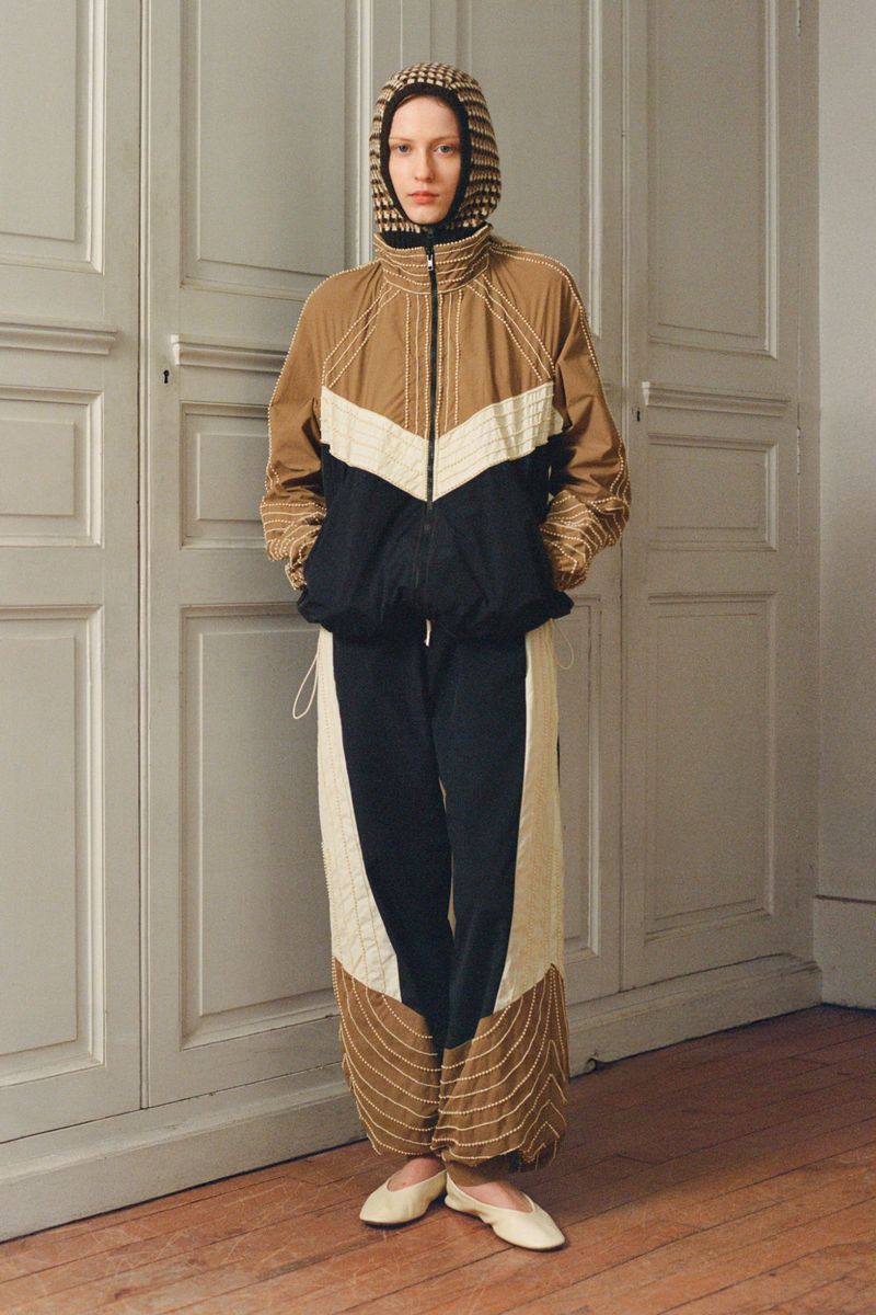 Fashion photo: A slender adult female model poses indoors wearing a brown, white, and black color-blocked hooded jacket and pants.