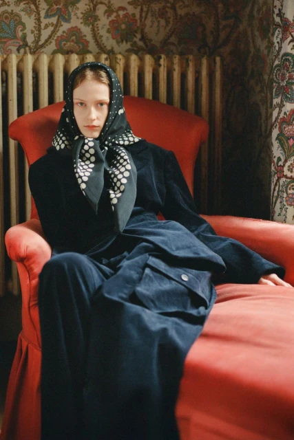 A woman in a navy blue outfit and polka dot headscarf sits in a red chair, indoors. The photo has a vintage editorial feel.