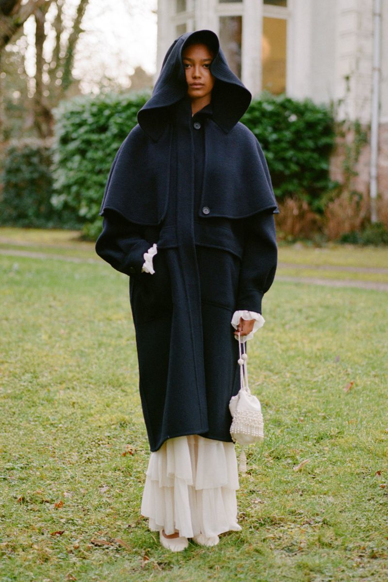 Woman in hooded black coat and ruffled-hem dress holds a beaded bag, posing outdoors on a grassy lawn with a building in the background.