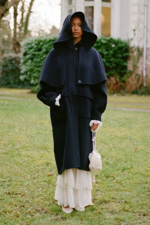 Woman in hooded black coat and ruffled-hem dress holds a beaded bag, posing outdoors on a grassy lawn with a building in the background.