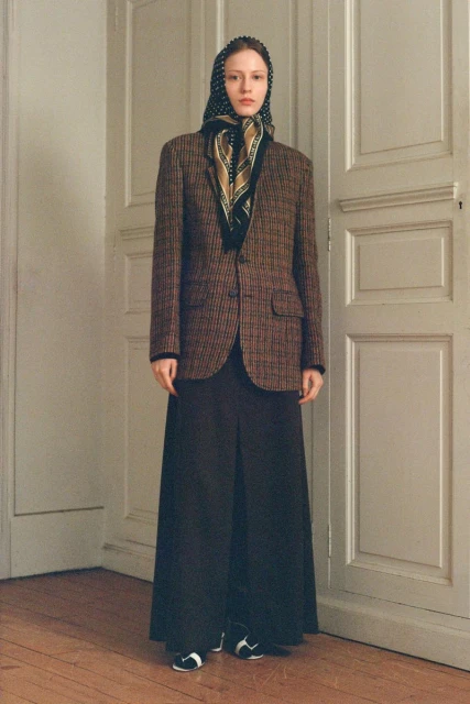An adult female poses indoors, wearing a brown tweed blazer, black pants, a patterned scarf and headscarf, standing against a white door.