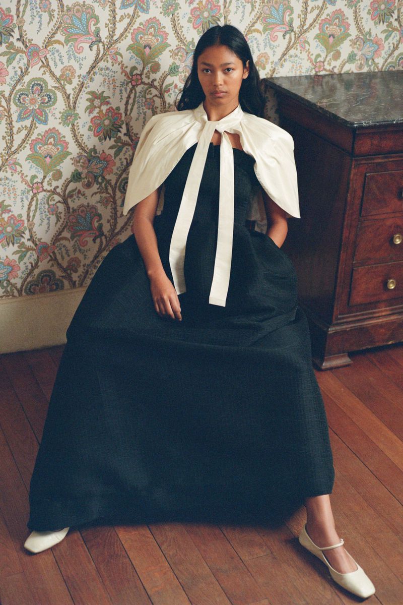 Elegant woman in a black dress with a white shawl and flat shoes sitting indoors with floral wallpaper and a wooden dresser.