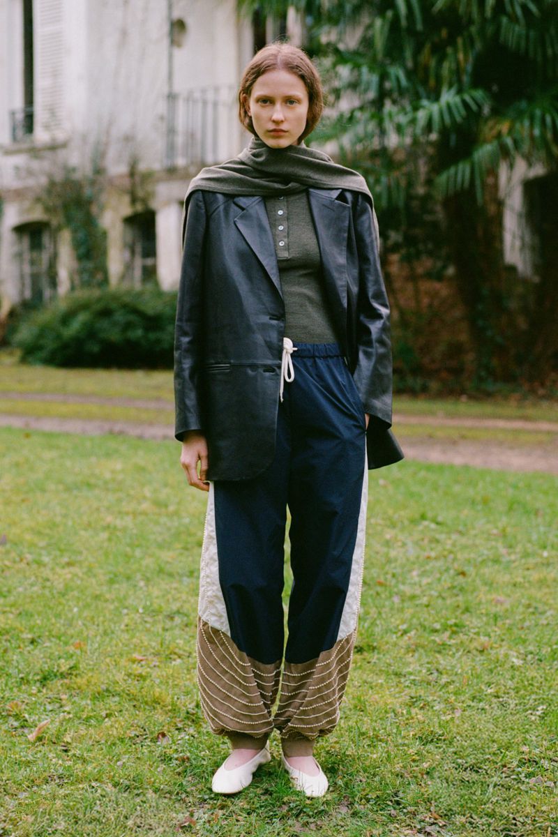 Full body fashion portrait of a woman wearing a blazer, scarf, and patterned pants standing in a park-like setting with a building.