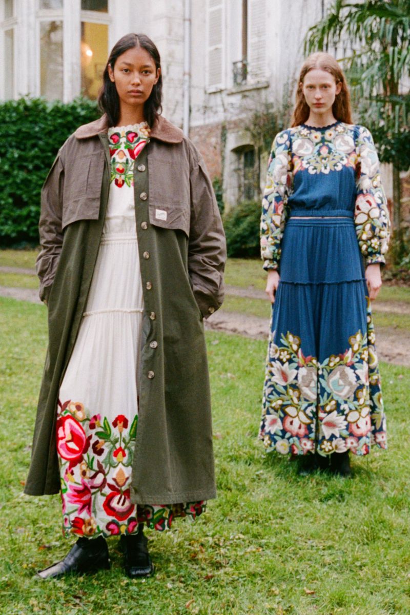 Two women model embroidered floral dresses and a trench coat in a lush outdoor garden setting against a vintage building backdrop.