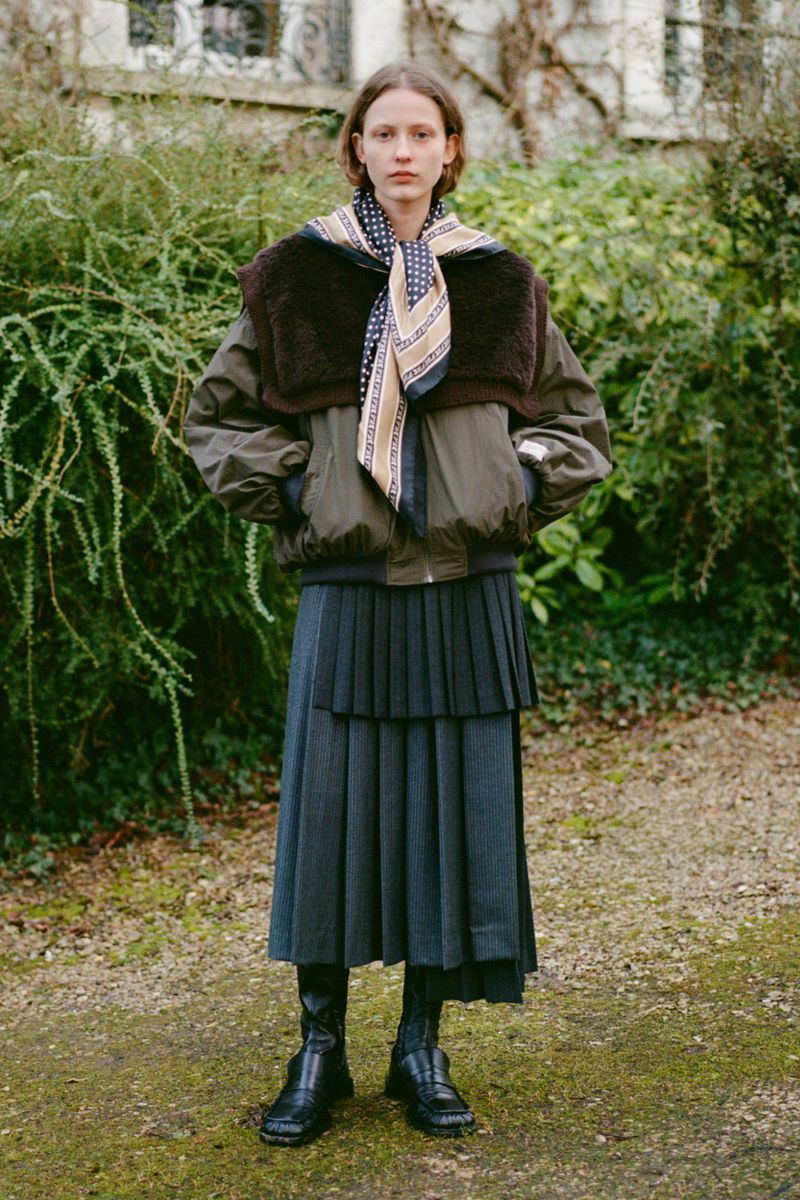 A woman stands in a garden wearing a green bomber jacket with a fur collar, layered skirt, patterned scarf and black boots.