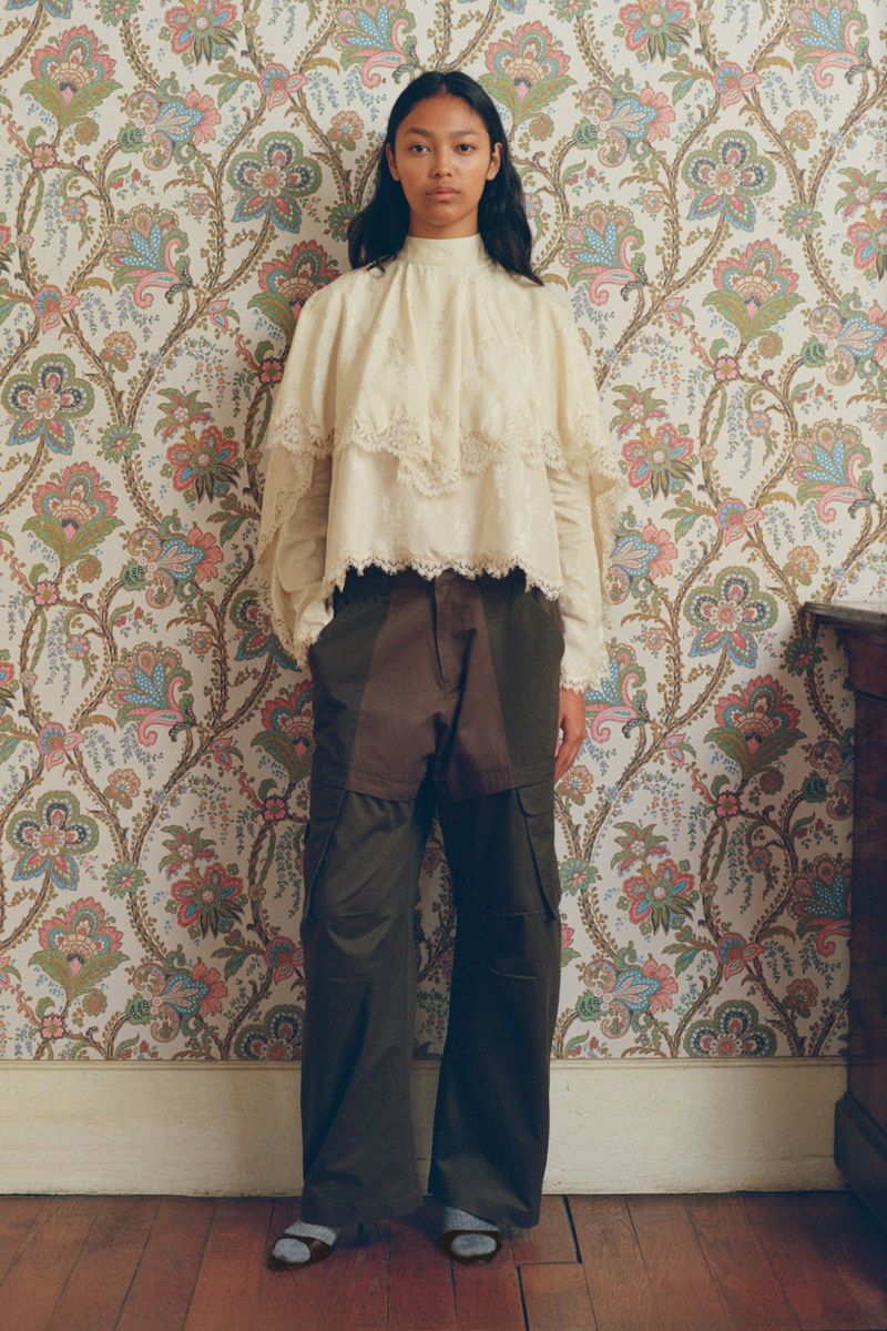 Woman in a cream lace blouse and dark pants stands against a patterned wallpaper. Fashion shot, indoor setting, natural light.