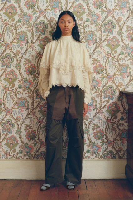Woman in a cream lace blouse and dark pants stands against a patterned wallpaper. Fashion shot, indoor setting, natural light.