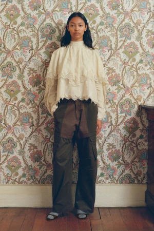 Woman in a cream lace blouse and dark pants stands against a patterned wallpaper. Fashion shot, indoor setting, natural light.