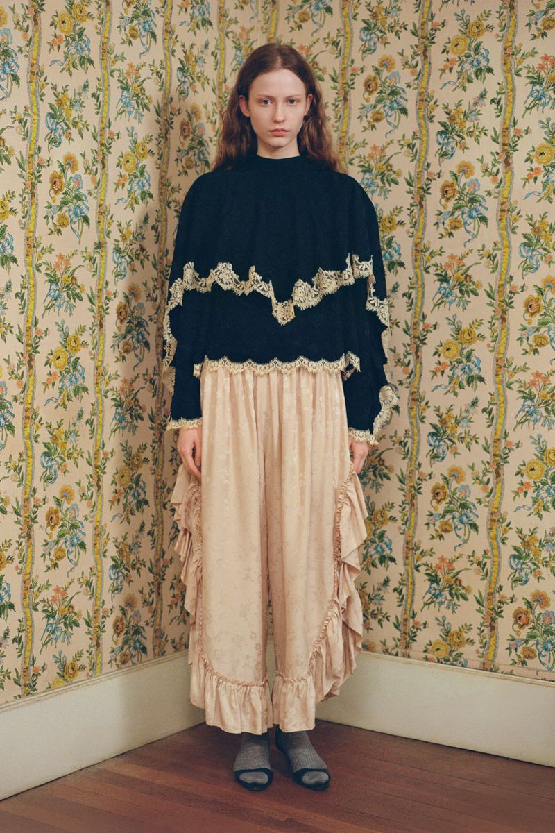 A young woman in a black top with lace trim and a ruffled skirt stands in a room with floral wallpaper, eye-level shot.