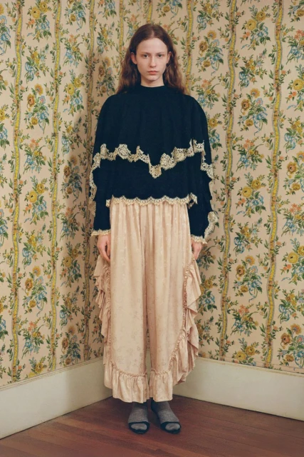A young woman in a black top with lace trim and a ruffled skirt stands in a room with floral wallpaper, eye-level shot.