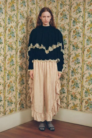 A young woman in a black top with lace trim and a ruffled skirt stands in a room with floral wallpaper, eye-level shot.