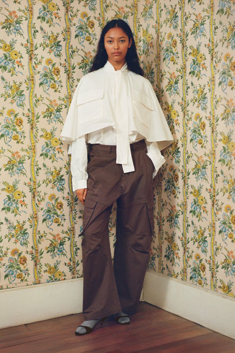 Full-length shot of a woman in a white cape blouse and brown cargo pants, posing in front of floral wallpaper. Fashion editorial.