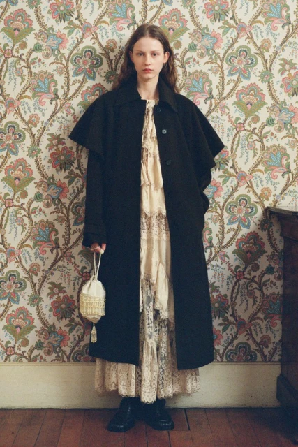 A full-length studio shot of a young female adult posing in a black coat and lace dress in front of floral wallpaper with a purse.