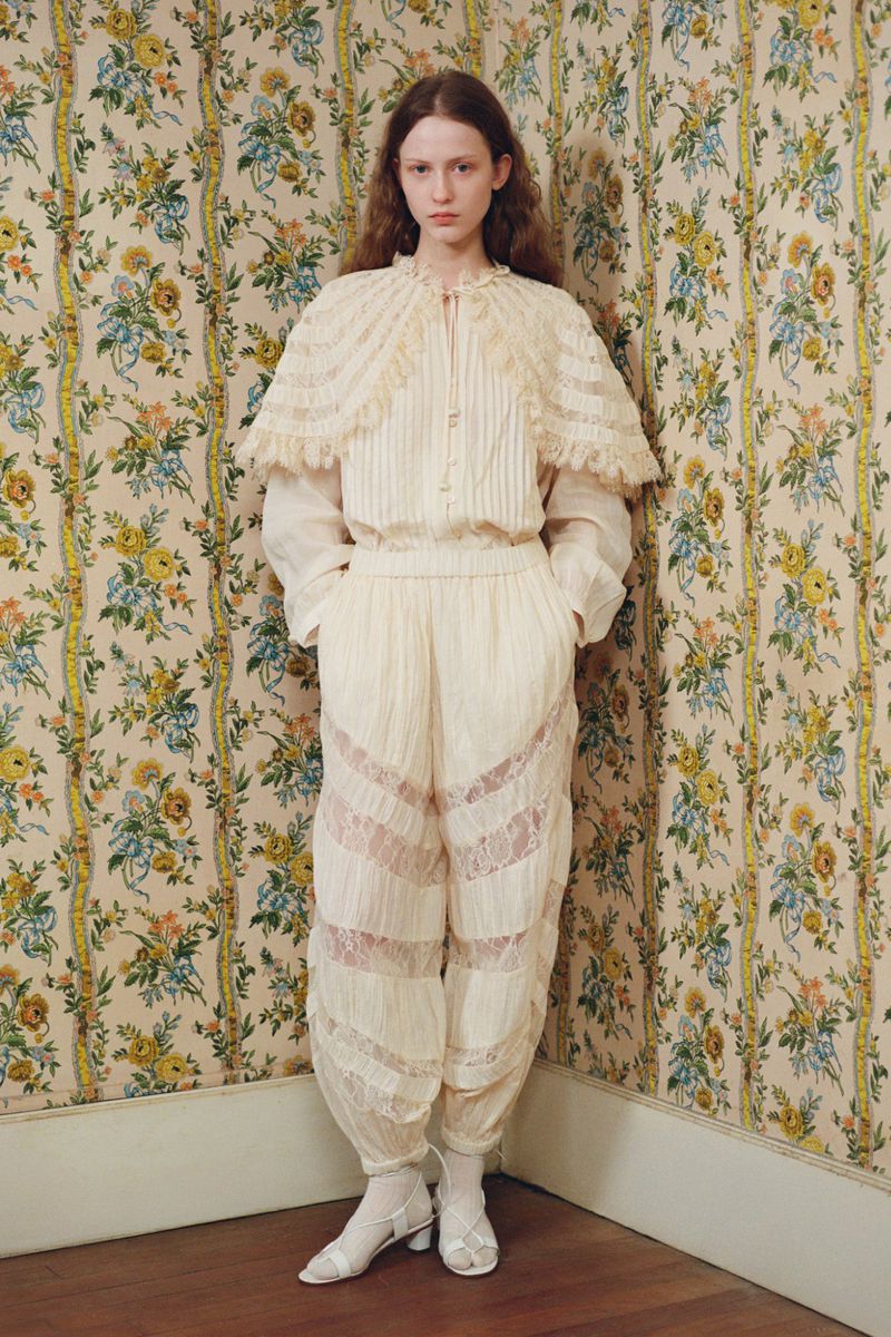 A fashionable adult female model poses indoors, wearing an ivory-colored, lacy outfit with floral wallpaper in the background.