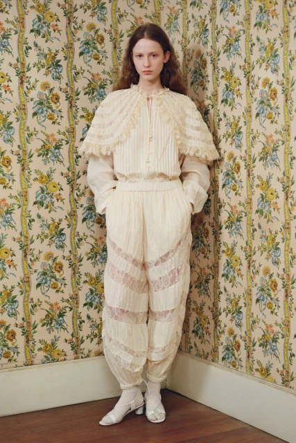 A fashionable adult female model poses indoors, wearing an ivory-colored, lacy outfit with floral wallpaper in the background.