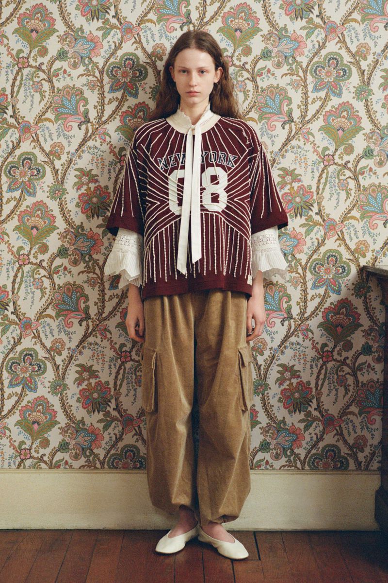 Portrait of a young woman in a maroon and white athletic-style shirt and baggy, golden-brown cargo pants, standing indoors against floral wallpaper.