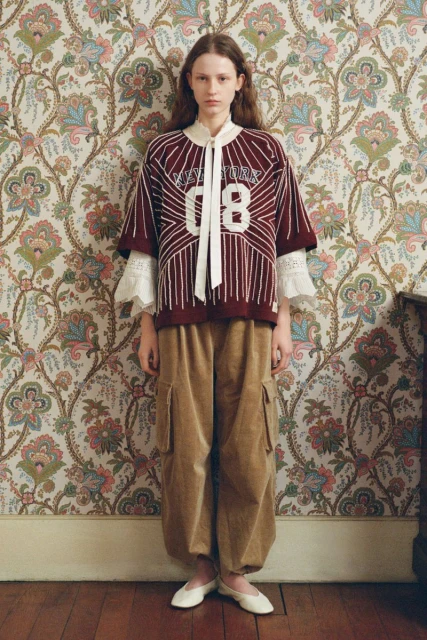 Portrait of a young woman in a maroon and white athletic-style shirt and baggy, golden-brown cargo pants, standing indoors against floral wallpaper.