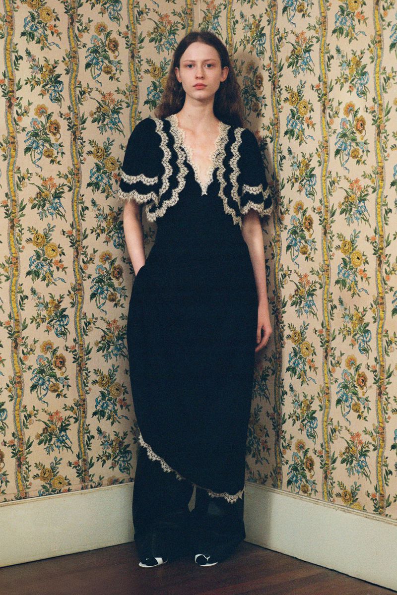 A pale-skinned woman in a black dress with lace trim stands in a corner of a room with floral wallpaper, posing with her hands in her pockets.