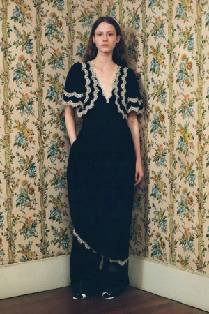 A pale-skinned woman in a black dress with lace trim stands in a corner of a room with floral wallpaper, posing with her hands in her pockets.