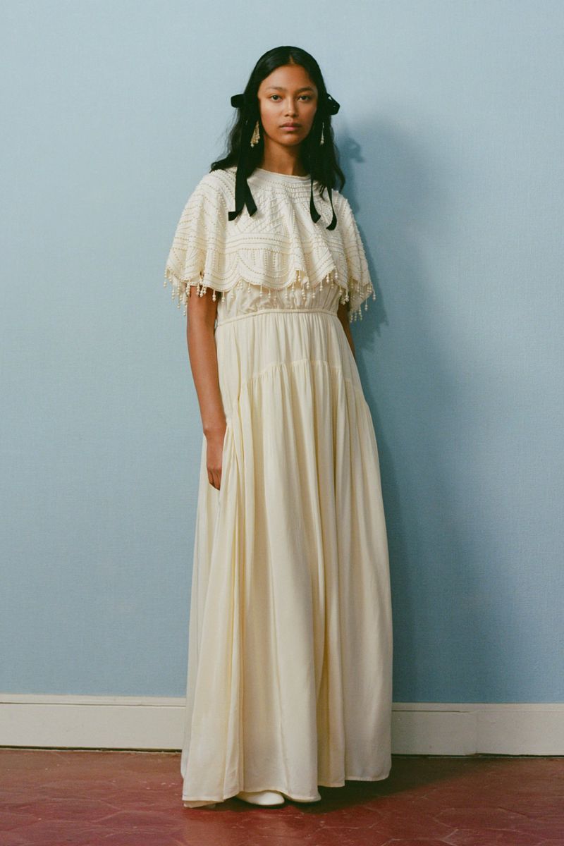 Full-length shot of a woman in a cream-colored dress with black hair ribbons, standing against a light blue wall. Studio lighting.