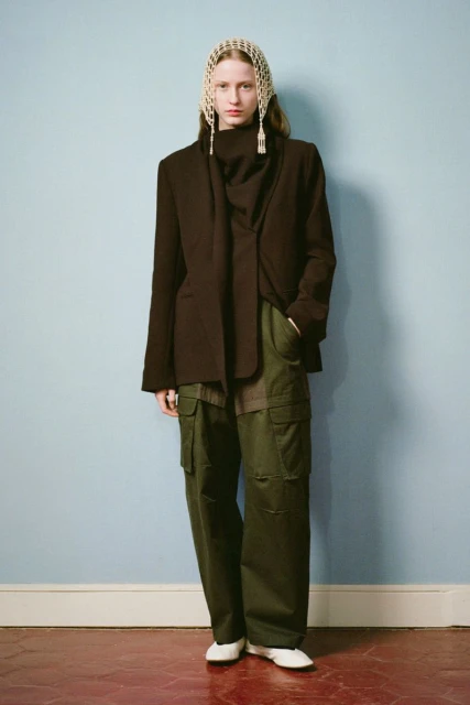 Full-length studio shot of a fashion model in an open brown blazer, green cargo pants, and a mesh head covering, against a blue wall.