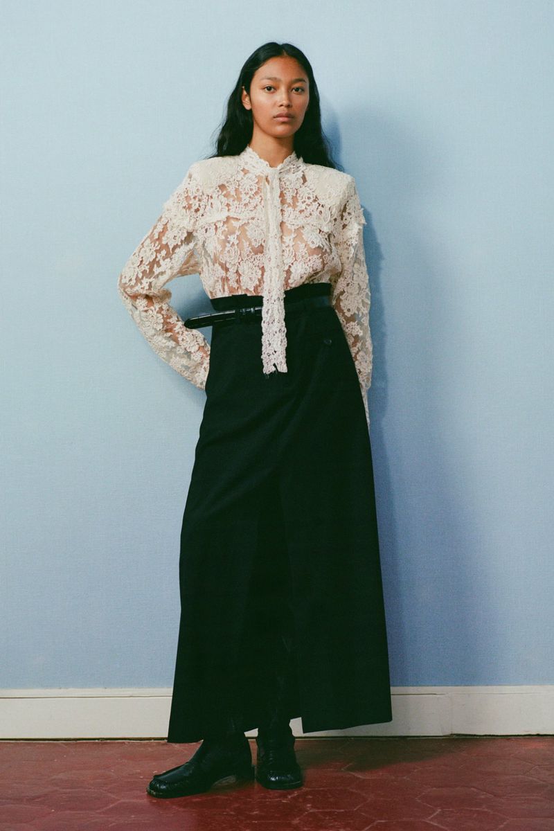 An adult woman poses in a studio wearing a white lace blouse with a tie, a black belt and long black skirt against a blue wall.