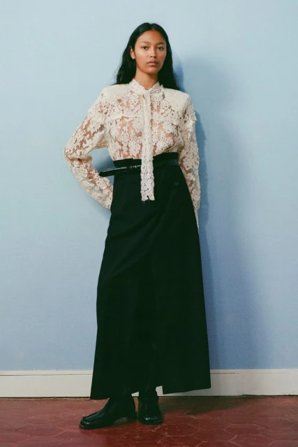 An adult woman poses in a studio wearing a white lace blouse with a tie, a black belt and long black skirt against a blue wall.