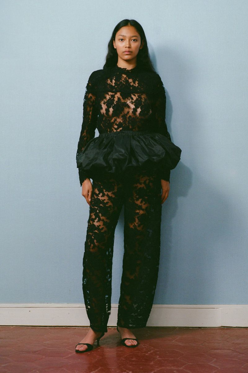 A slender adult woman in a high-fashion black lace outfit poses against a light-blue wall, emphasizing style and texture, in a studio setting.
