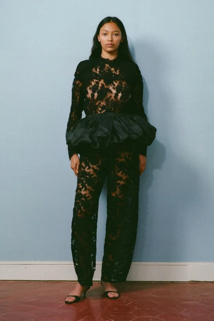 A slender adult woman in a high-fashion black lace outfit poses against a light-blue wall, emphasizing style and texture, in a studio setting.