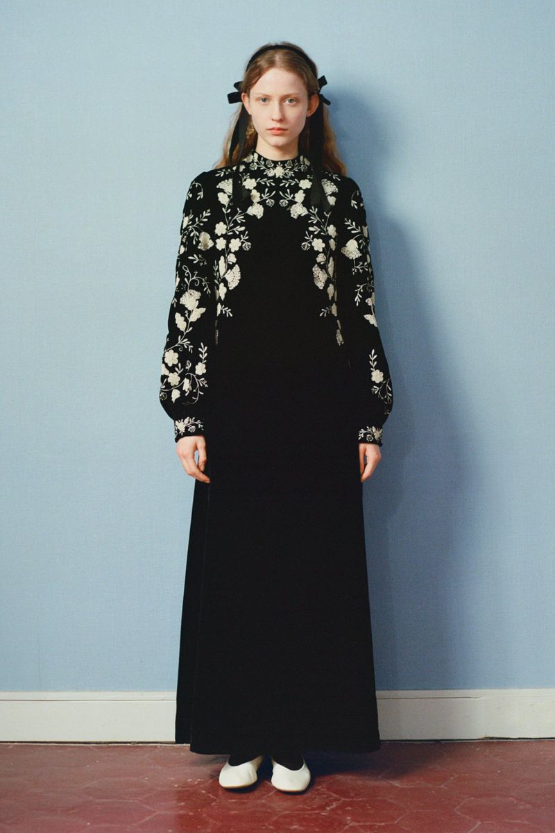 Full shot of a red-haired woman in a black dress with floral embroidery posing against a blue wall in a studio. Fashion editorial.