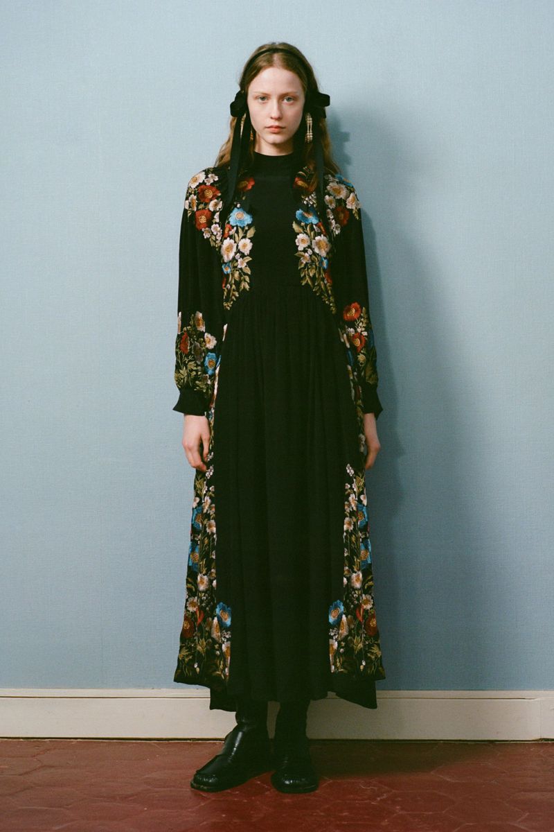A young woman stands in a photo studio wearing a long black dress with floral embroidery and matching black boots and hair ribbons.