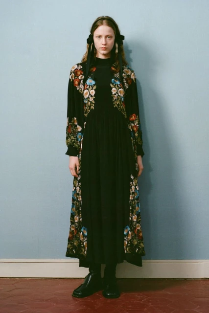 A young woman stands in a photo studio wearing a long black dress with floral embroidery and matching black boots and hair ribbons.
