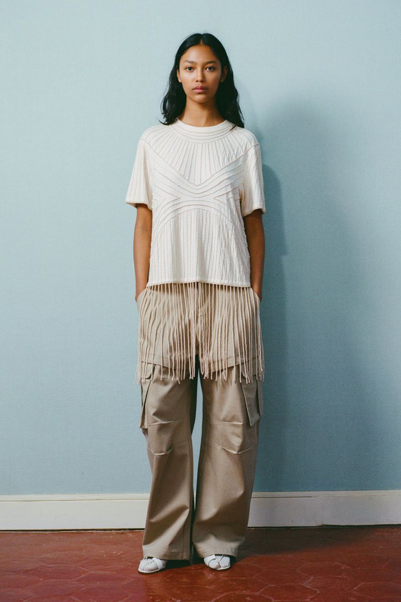 A woman poses in a cream-colored t-shirt and khaki cargo pants with a light blue background. She is standing with her hands in her pockets.