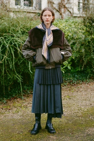Model wearing a brown bomber jacket, patterned scarf, and pleated midi skirt outdoors in a garden